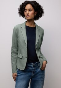 Femme aux cheveux bouclés portant un blazer vert texturé, une chemise bleu marine et un jean bleu, debout avec une main dans la poche devant un fond uni.