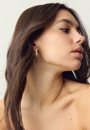 Nuori nainen, jolla on pitkät tummat hiukset, ja kultaiset rengaskorvakorut, joissa on pienet valkoiset timantit, katsomassa oikealle, paljaat hartiat näkyvissä.