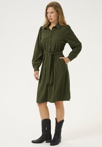 Robe en velours côtelé vert avec manches longues, boutonnage à l'avant et taille nouée. Porte avec des bottes de cowboy noires, dotées de coutures décoratives.