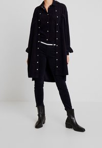 Persona che indossa un lungo cappotto nero con bottoni, pantaloni neri, una camicia nera, una cintura bianca e stivali neri alla caviglia, in piedi contro uno sfondo semplice.