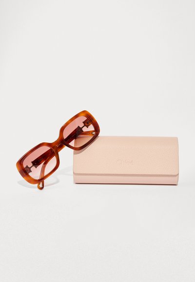 Gafas de sol de gran tamaño de carey con lentes rosas, acompañadas de un estuche texturizado rosa con un logo grabado en la parte frontal.