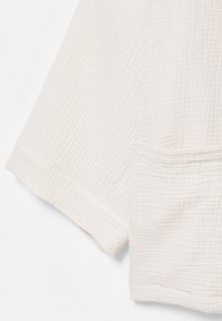 Tissu blanc texturé avec un motif de grille subtil, montrant une couture d’angle et un petit détail de poche.