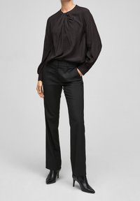 Blusa nera, con texture, e scollatura rotonda, con dettaglio arricciato al centro, abbinata a pantaloni neri su misura e décolleté neri a punta.