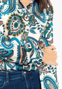 Person som bär en skjorta med paisleymönster i blått, grönt och brunt tillsammans med mörkblå denimjeans, med armarna i kors över kroppen.