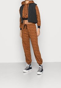 Tuta marrone a fantasia con un top nero corto. Include un cappuccio e pantaloni con coulisse, abbinati a sneaker nere alte.