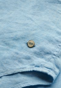 Lichtblauw linnen shirt met een gladde textuur, voorzien van een ronde gouden speld met wit logo en fijne stiksels langs de randen.