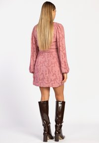 Roze jurk met bloemenpatroon, lange mouwen, een getailleerde taille, en glanzende bruine laarzen tot de knie, geshowd door een model van achteren.
