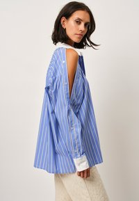 Blue striped shirt with a white collar and cuffs, featuring cut-out shoulders, long sleeves, and button details, fabric appears smooth.