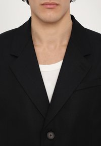 Schwarzes Blazer aus einem strukturierten Stoff, mit einem Reverskragen, einem einzelnen Knopfverschluss und einer Brusttasche. Getragen über einem weißen Unterhemd.