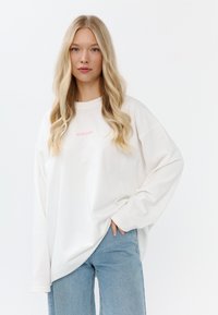 Witte oversized sweatshirt met lange mouwen en roze tekst "MIRONS" op de borst, gecombineerd met lichtblauwe denim jeans. Soepele textuur.