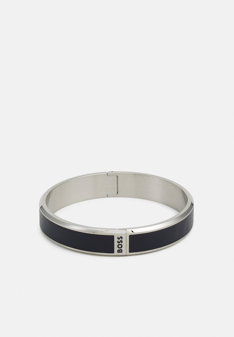 Bracelet en métal argenté avec une bande en cuir noir. Présente un design lisse et arrondi avec le logo "BOSS" sur un accent métallique.