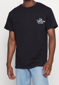 Czarny bawełniany t-shirt z krótkimi rękawami i okrągłym dekoltem. Na lewej piersi nadrukowany biały tekst "ON VACATION CLUB".