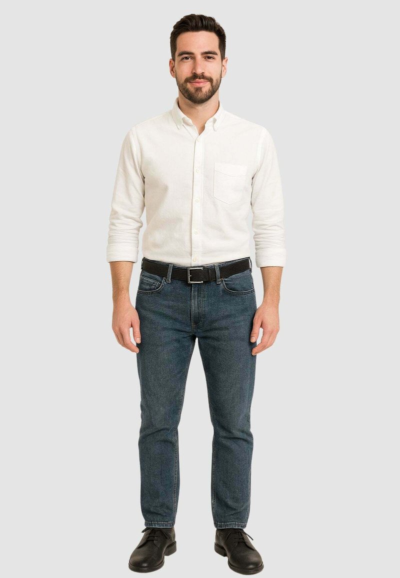 Camicia bianca a maniche lunghe con bottoni, con taschino sul petto, abbinata a jeans blu scuro e scarpe nere. Design semplice e aderente con colori classici.