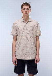 Beige pikétröja med bladmönster i brunt, kortärmad och med krage. Bärs med mörkblå shorts.