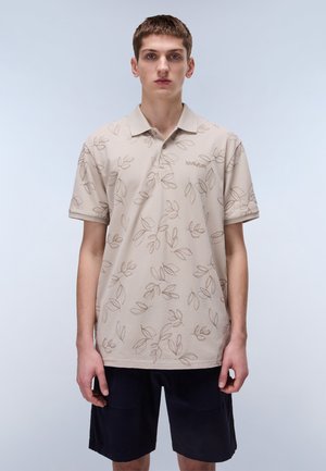 Beige poloshirt med bladmønstre i brun, korte ærmer og krave. Båret med mørkeblå shorts.