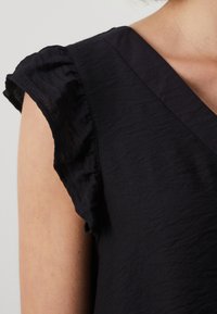 Musta mekko, jossa on lyhyet röyhelöhihat ja joka on valmistettu teksturoidusta kankaasta. Mekossa on V-kaula-aukko ja sileä, kevyt materiaali.