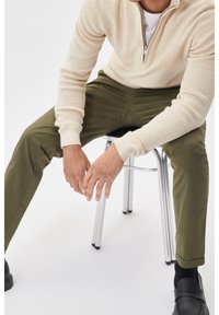 Beige knit zip-up sweater, green chinos, and black loafers. Model seated on a silver chair, hands resting on knees. Simple and clean design.