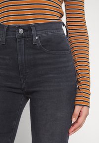 Pantalones vaqueros negros de cintura alta con una textura suave, cierre de botón metálico y diseño de cinco bolsillos, combinados con una blusa de manga larga a rayas en tonos óxido y negro.