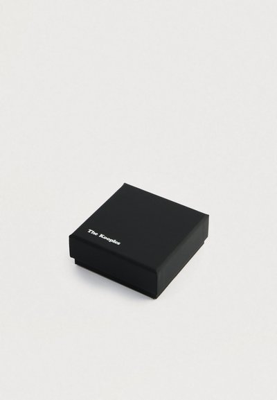 Pequeña caja cuadrada negra con el logo blanco de "The Kooples" en la parte superior, sobre un fondo claro y liso.