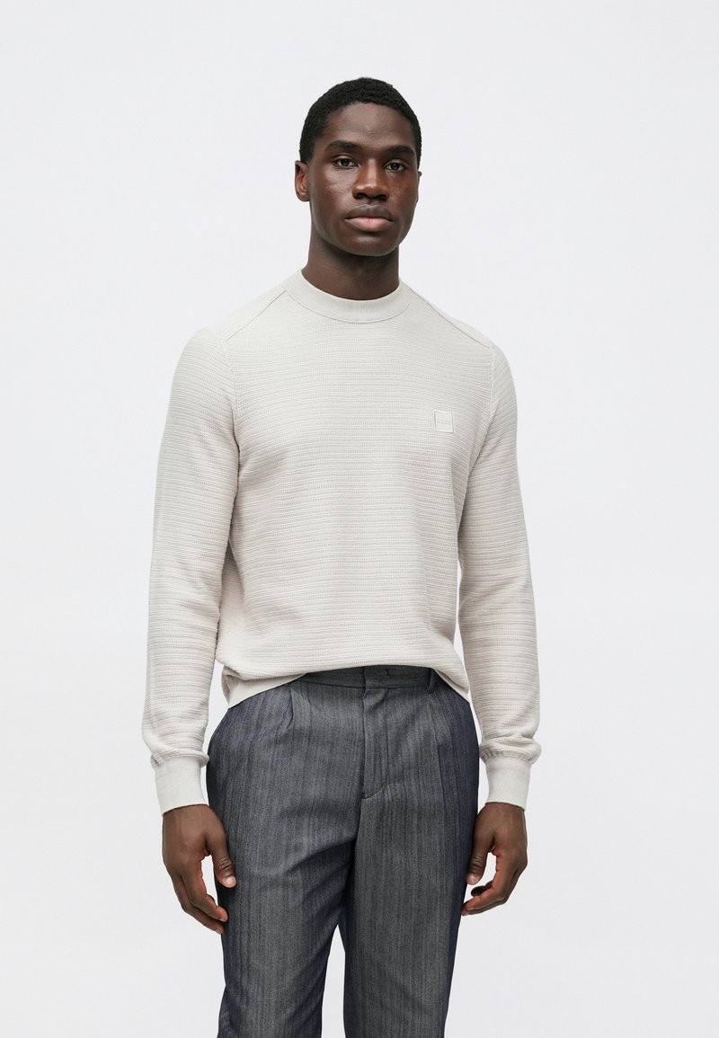 Gaiši bēšs adīts džemperis ar riboto faktūru un apaļu kakla izgriezumu, kombinēts ar pelēkām svītrainām biksēm. Uz krūtīm ir neliela zīmola etiķete.