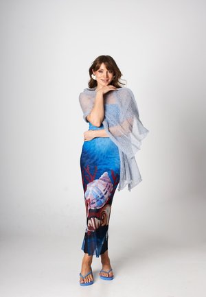 Kobieta uśmiechająca się, ubrana w niebieską sukienkę o tematyce morskiej z muszlami, jasnoszary szal, kolczyk w kształcie kwiatu i niebieskie sandały na jednolitym tle.