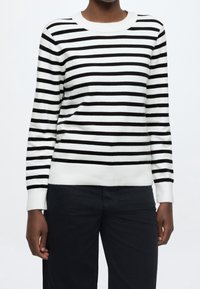 Personne portant un pull blanc avec des rayures horizontales noires et un pantalon noir, debout devant un fond clair uni.