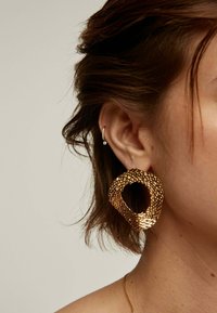 Pendiente de aro texturizado en oro con un diseño abierto, que presenta un patrón trenzado y un acabado pulido, llevado en la oreja.