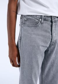 Vaqueros de denim gris con una textura desgastada, diseño de cinco bolsillos, costuras visibles y herrajes metálicos, combinados con una camiseta blanca.