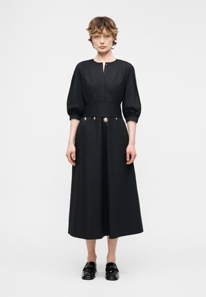 Femme debout de face, portant une robe midi noire avec des manches bouffantes, des breloques décoratives sur la ceinture, et des mocassins noirs, sur un fond uni.