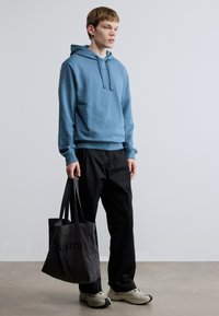 Sweatshirt azul com capuz, cordão e punhos canelados, combinada com calças pretas. Uma mala tote cinzenta pendurada na mão, com texto impresso.
