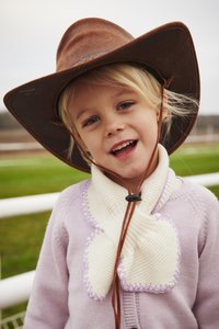 Brūna plata plata kovboju cepure, gaiši violeta džemperis ar adītu apkakli un krēmkrāsas kaklasaits ar lavandas šuvēm un pogas aizdari.