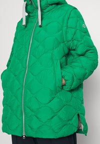 Veste matelassée verte en doudoune avec capuche, dotée d'une fermeture éclair à l'avant, de deux poches latérales et de cordons grijs. Surface douce et texturée.