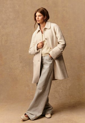 Femme portant un manteau en laine crème sur un pull en tricot, un pantalon large gris clair et des chaussures beiges, se tenant contre un fond marron neutre.