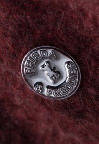 Donker burgundy stof met een zachte textuur heeft een ronde zilveren metalen badge. De badge toont "FRIEDA & FREDDIES" en een kleine hondensilhouet.