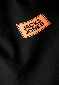 Черен плат с оранжева правоъгълна лепенка, показваща логото "JACK & JONES" с дебели черни букви.