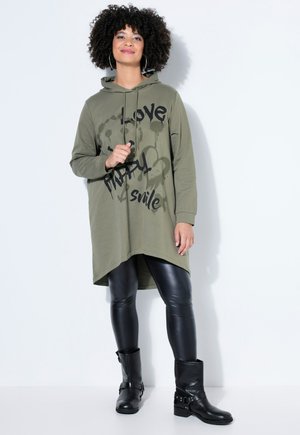 Frau mit lockigem Haar, die ein olivgrünes Hoodie-Kleid mit dem Schriftzug "Love Happy Smile", schwarze Lederleggings und schwarze Stiefeletten trägt.