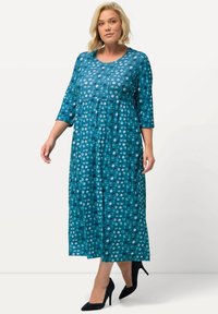 Teal mekko, jossa on kolme neljäsosaa hihat, pyöreä kaula-aukko ja kootut vyötärö. Koko mekkoa koristaa monivärinen pistekuvio. Vaatteena mustat korot.