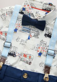 Tenue pour enfants avec une chemise blanche ornée d’illustrations colorées de villes et d’animaux, un nœud papillon bleu marine, des bretelles bleu clair et un pantalon bleu marine avec des boutons en bois.