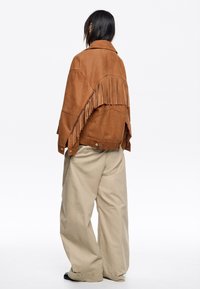 Bruine suède jas met franjes, voorzien van een brede kraag en drukknoopsluiting. Gecombineerd met losse beige broek en zwarte laarzen.
