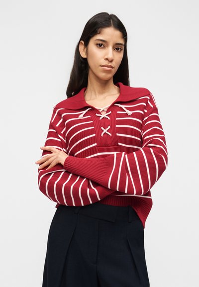 Γυναίκα με μακριά σκούρα μαλλιά, ντυμένη με πουλόβερ με κόκκινες και λευκές ρίγες, με κορδόνια στον λαιμό, και μαύρο παντελόνι, στέκεται με σταυρωμένα τα χέρια.