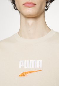 Beige katoenen sweatshirt met een witte geborduurde "PUMA"-logo en een oranje accent ontwerp eronder. Standaard ronde halslijn en ribgebreide manchetten.