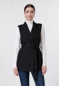 Μια νεαρή γυναίκα με καστανά μαλλιά φοράει ένα αμάνικο μαύρο σακάκι με ζώνη πάνω από ένα λευκό πουλόβερ ζιβάγκο και μπλε τζιν, στέκεται μπροστά σε λευκό φόντο.