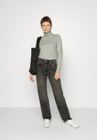 Grauer Rollkragenpullover, hoch taillierte schwarze Jeans mit fader Textur und weiße Sneaker. Schwarze gesteppte Schultertasche. Schlicher Hintergrund.