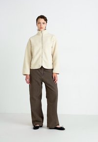 Cream-colored fleece jacket with a high collar, front zipper, and textured pattern, paired with loose brown trousers and black flat shoes.