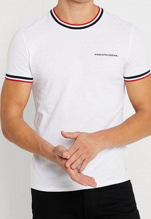 Man draagt een wit T-shirt met rode en zwarte bies op kraag en mouwen, handen voor zich samengevouwen, tekst "MARCOPOLODENIM" op de borst.