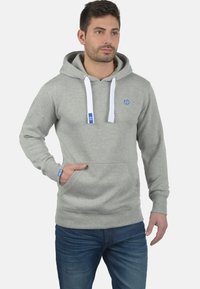 Siva dukserica od mješavine pamuka, s prednjim džepom, bijelim vezicama i plavim logotipom na prsima i manžetama. Uparena s plavim trapericama.