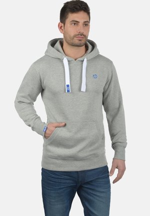 Siva dukserica od mješavine pamuka, s prednjim džepom, bijelim vezicama i plavim logotipom na prsima i manžetama. Uparena s plavim trapericama.