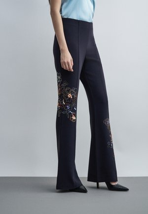 Γυναίκα που φορά μαύρο καμπάνα παντελόνι με πολύχρωμη κεντητή λουλούδια στο πλάι, συνδυασμένο με μαύρες μυτερές γόβες και κοντό γαλάζιο τοπ.