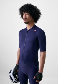 Dunkelblaues Fahrradtrikot mit kurzen Ärmeln, Reißverschluss vorne und strukturiertem Material. Getragen mit passenden Bib-Shorts und einem weißen Helm in der Hand.