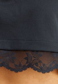 Dobladillo de tela oscura con borde de encaje floral negro que muestra finos patrones de malla y hojas sobre fondo beige.